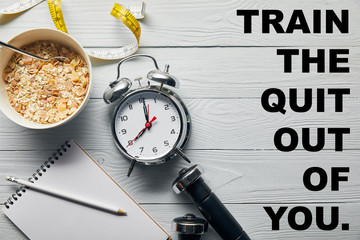 top view of silver alarm clock, notebook with pencil, breakfast cereal, measuring tape, dumbbells on wooden white background with train the quit out of you lettering