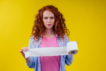 Beautiful curly redhead ginger woman holding toilet paper roll on yellow color background