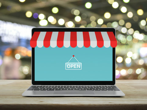 Modern Laptop Computer With Online Shopping Store Graphic And Open Sign On Wooden Table Over Blur Light And Shadow Of Shopping Mall, Business Internet Shop Online Concept