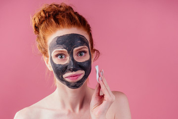 redhaired ginger teenager girl with black clay mask on her pretty face on pink studio background