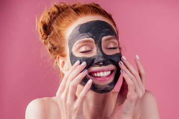 redhaired ginger teenager girl with black clay mask on her pretty face on pink studio background