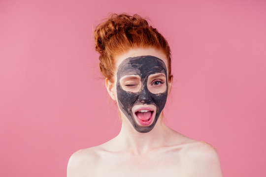 redhaired ginger teenager girl with black clay mask on her pretty face on pink studio background