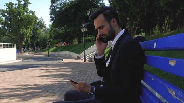 businessman using a mobile phone in a park on a bench. man talking on a cell phone. bearded man talking on a smartphone. young attractive bearded man is sitting and talking on a cell phone with a frie