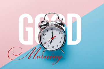top view of classic silver alarm clock on pink and blue background with good morning lettering