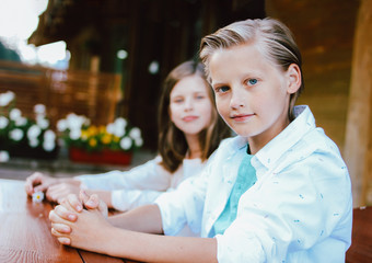 Attractive preteen blonde boy with blue eyes in white shirt with sister friend sitting at table in backyard of wooden cottage © Galina Zhigalova