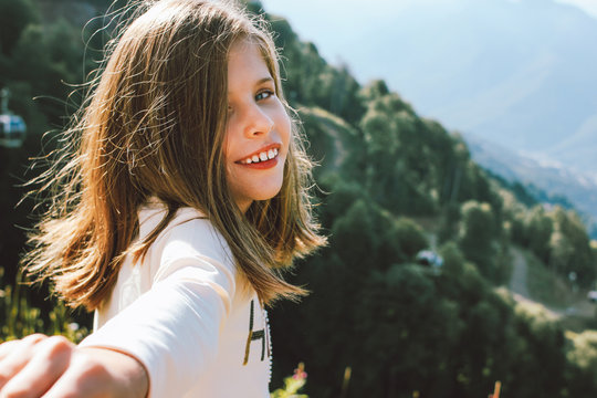 Smiling tween girl reaches out to camera follow me on background of beautiful mountains, family travel concept