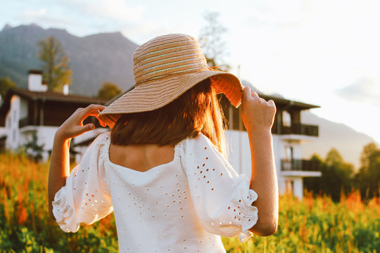 Beautiful romantic preteen girl in straw hat against background of beautiful houses in mountain, rural scene at sunset. View from behind, golden hour