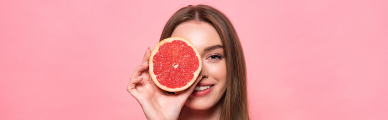 panoramic shot of smiling attractive girl holding cut grapefruit isolated on pink