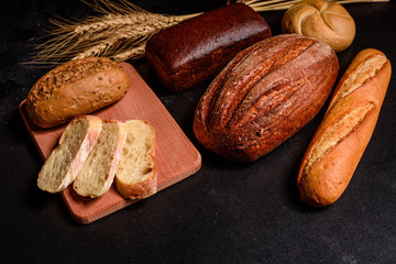 Obraz premium Fresh fragrant bread with grains and cones of wheat against a dark background. Assortment of baked bread on wooden table background. Fresh fragrant bread on the table. Food concept. 