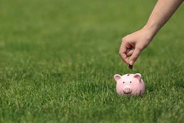 Young woman putting coin into piggy bank on green grass outdoors, closeup. Space for text