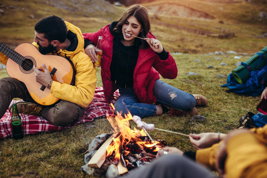 Group of smiling friends with marshmallow sitting around bonfire in camping