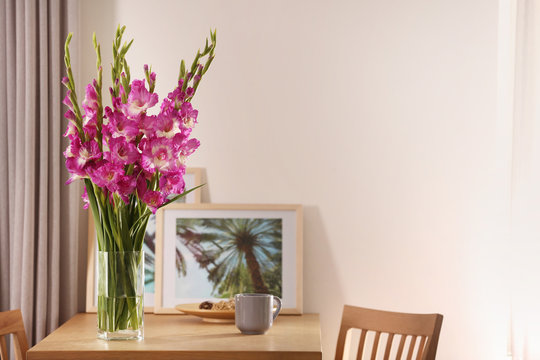 Vase with beautiful pink gladiolus flowers, pictures and cup on wooden table in room, space for text