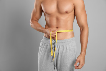 Young man with slim body using measuring tape on grey background, closeup view
