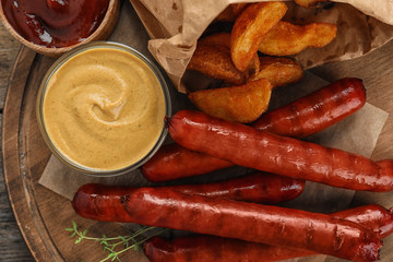 Delicious grilled sausages with fried potatoes and sauces on wooden board, closeup. Barbecue food