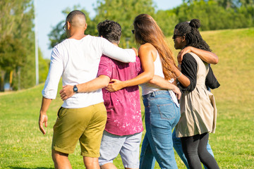Back view of friends hugging while strolling on meadow. Young people talking while walking together. Friendship concept
