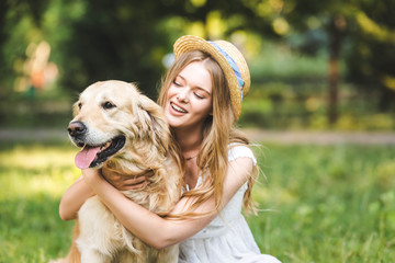 selective focus of beautiful girl in white dress and straw hat hugging golden retriever and smiling while sitting on meadow