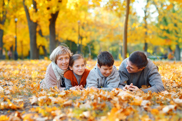 Naklejka premium Happy family lying on fallen leaves, playing and having fun in autumn city park. Children and parents together having a nice day. Bright sunlight and yellow leaves on trees, fall season.