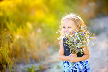 lovely cute little girl with blond hair walks outdoors at sunset, holds a bouquet of wildflowers in...