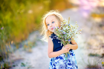 lovely cute little girl with blond hair walks outdoors at sunset, holds a bouquet of wildflowers in...