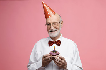 Cheerful stylish bearded man in his eighties wearing red cone party hat, holding piece of delicious...