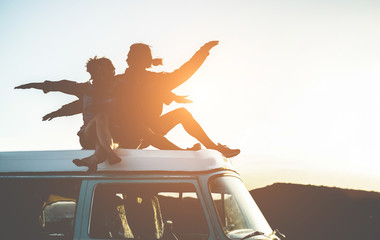 Happy couple silhouette sitting on top of minivan at sunset - Young people having fun on summer...