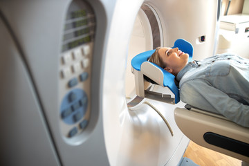 Smiling young woman lying on CT machine table at radiology clinic - obrazy, fototapety, plakaty