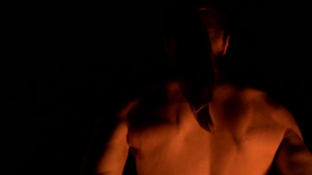 Close-up back view. Low key. Young male with long hair and bare torso rotates burning torch outdoors on a black night video slow motion. Modern fakir does tricks with a burning staff