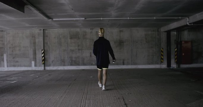 TRACKING Teenager girl soccer player walking towards a wall in empty covered parking garage, holding spray paint can in her hands. 4K UHD 60 FPS RAW graded footage