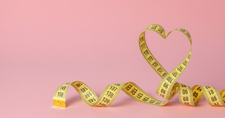 Yellow measuring tape in the shape of a heart on a pink background. The concept of weight loss for the normal functioning of the heart and body. Copy space for text.