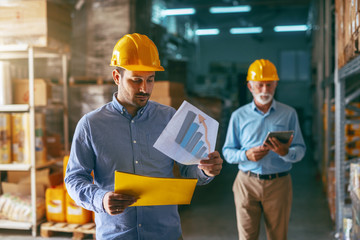 Supervisor holding folder with charts and analyzing sales while standing in warehouse. In background older warehouse man using tablet. Warehouse interior.