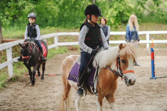 Children with helmets and protective vests on riding pony horses at sunny day on ranch.