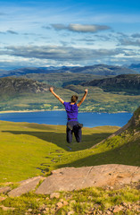 Young Boy Jumps Happily On Rock In Front Of Spectacular Landscape Of Applecross Pass In Scotland
