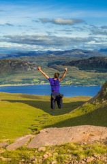 Fototapeta premium Young Boy Jumps Happily On Rock In Front Of Spectacular Landscape Of Applecross Pass In Scotland