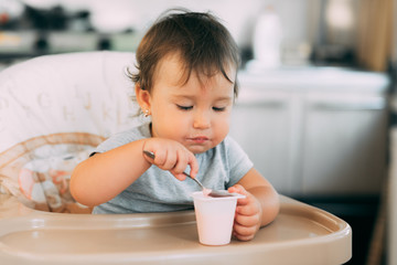 Cute, little girl eating yogurt in the kitchen during the day in a high chair