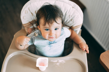 Cute, little girl eating yogurt in the kitchen during the day in a high chair