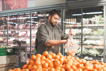 Happy man with a beard stands in a supermarket in the fruit department and puts tangerines in a package. Smiling buyer selects tangerines in a grocery store. Shopping in a supermarket.
