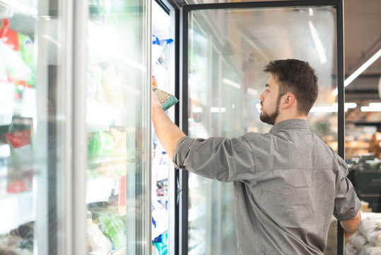 Man in his shirt takes his hand frozen foods from the refrigerator in the supermarket. The buyer selects the products in the grocery store's refrigerator.