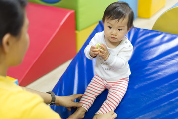 Young little Asian baby holding toy ball playing with female preschool teacher in classroom. Kid...