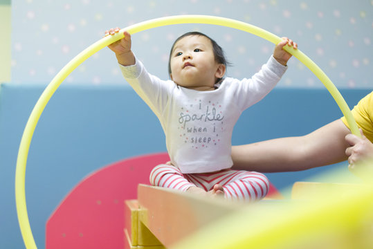 Young little Asian baby holding toy loop playing with preschool teacher in baby play gym. Kid enjoy learning and discover new things in the class while nanny holding her. Play and learn concept.