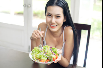 Young good health beautiful smiling Asian long hair girl is eating healthy food such as green salad sitting at home. She is looking at cabbage on her fork. Healthy food and diet concept.