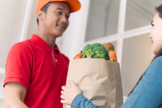 Asian Delivery Man In Red Color Uniform Handling Bag Of Foods Or Vegetables To Young Beautiful Female Costumer In Front Of The House. Postman Or Express Delivery Service Concept. Focus On Girl Eyes.