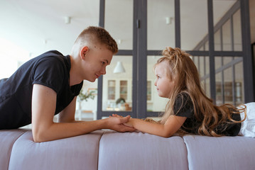 caucasian siblings - teenager boy brother and little girl sister laying holding hands on a couch (sofa) in modern loft interior