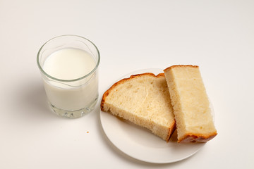 Glass of milk and two pieces of white bread on the white saucer are on the white background/table. International Day to Assist the Poor	