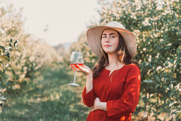 Wine tasting and relaxing in the garden. Young beautiful woman in red dress and hat, gracefully holding a glass of wine. There are fruit trees in the background