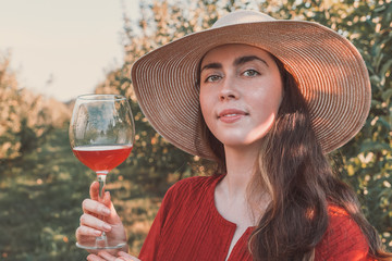 Wine tasting and relaxing in the garden. Young beautiful brunette full face in a red dress and hat with a glass of wine. There are fruit trees in the background. Tint