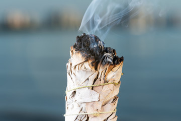 Smudging Ritual using burning thick leafy bundle of White Sage on the beach at sunrise in front of the lake.