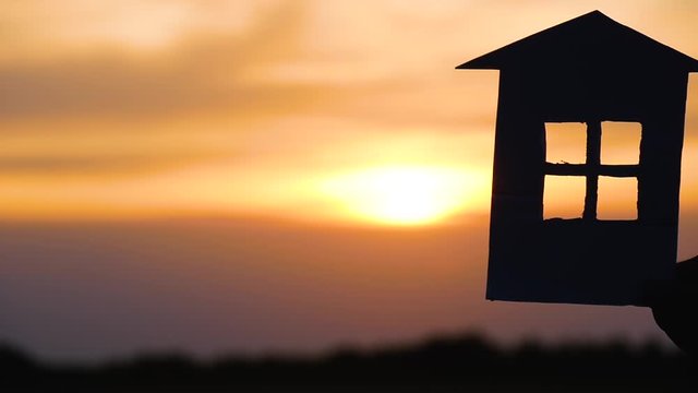 The silhouette of the house from paper on background of sunset. A man's hand holds a model of a paper house. The concept of construction and housing lending.