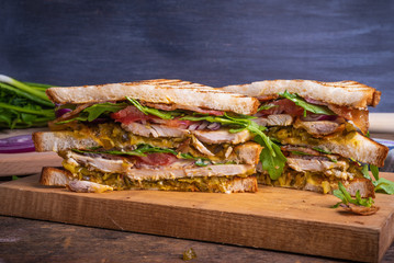 Delicious sandwich cut into triangles - toasted bread with chicken breast, fried bacon, vegetables, pickles with mustard and arugula closeup on a dark background, copy space