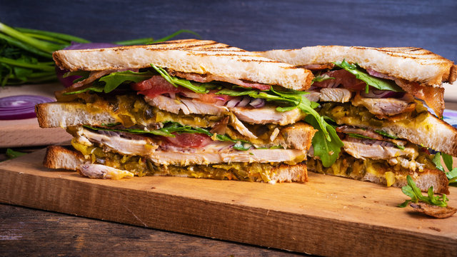 Delicious sandwich cut into triangles - toasted bread with chicken breast, fried bacon, vegetables, pickles with mustard and arugula closeup on a dark background, copy space