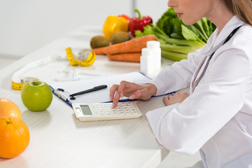 cropped view of dietitian in white coat using calculator at workplace
